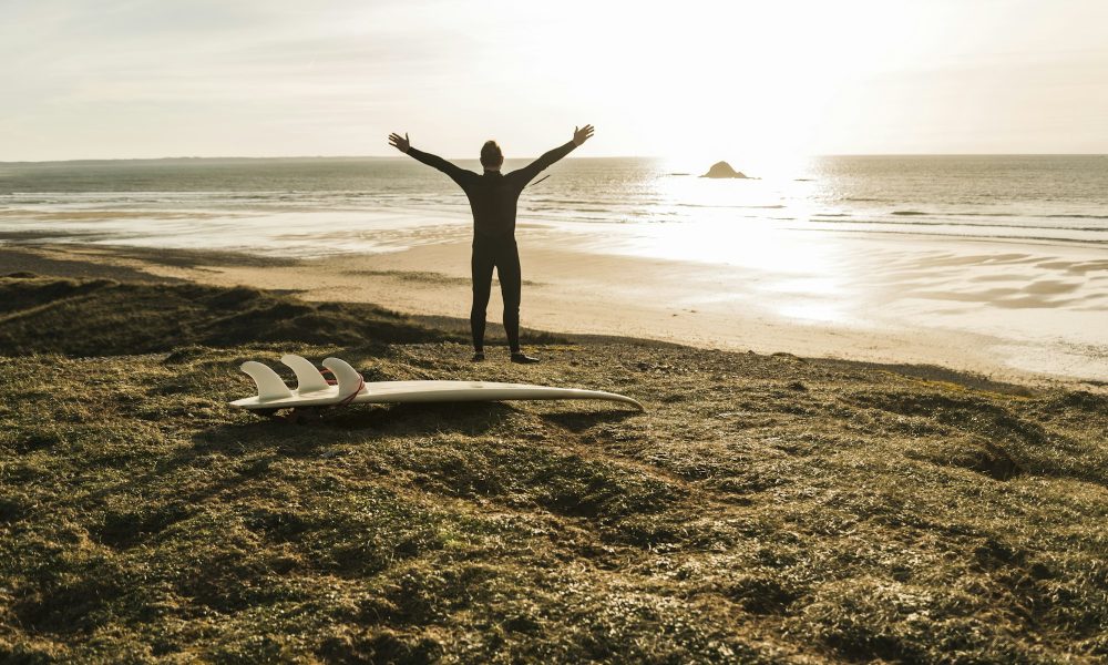 France, Bretagne, Finistere, Crozon peninsula, man at the coast with outstretched arms and surfboard