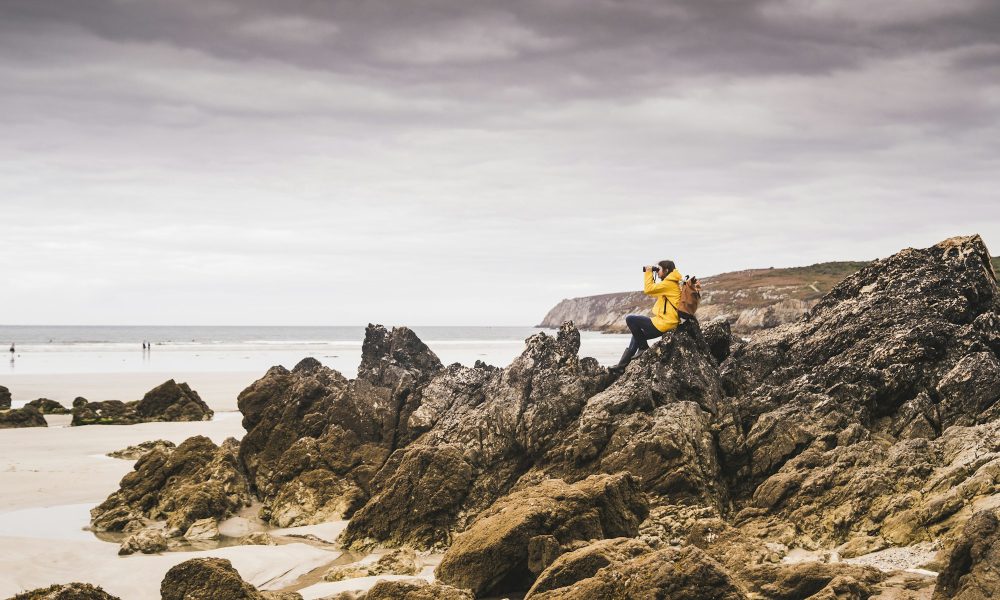 Young woman wearing yellow rain jacket at the beach, looking through binoculars, Bretagne, France